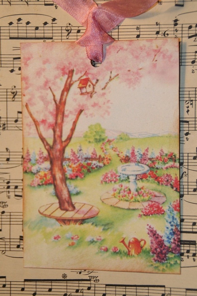 May include: A vintage-style illustration of a garden scene with a pink flowering tree, a birdhouse, a birdbath, and a watering can. The illustration is printed on a tag with a pink ribbon.
