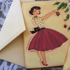 Op de afbeelding: Vintage kerstkaart met een vrouw in een top met stippen en een rode rok die een kerstboom versiert. De kaart zit in een cr&egrave;me-kleurige envelop. De achtergrond is een witte kanten stof.