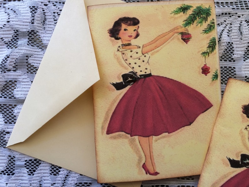 Op de afbeelding: Vintage kerstkaart met een vrouw in een top met stippen en een rode rok die een kerstboom versiert. De kaart zit in een cr&egrave;me-kleurige envelop. De achtergrond is een witte kanten stof.