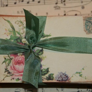 May include: A stack of vintage-style envelopes tied with a green ribbon and decorated with floral illustrations. The envelopes have a faded, antique look and feature a floral design with a pink rose, purple flowers, and green leaves.