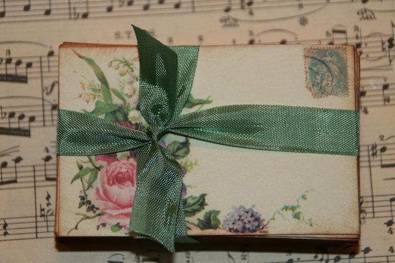 May include: A stack of vintage-style envelopes tied with a green ribbon and decorated with floral illustrations. The envelopes have a faded, antique look and feature a floral design with a pink rose, purple flowers, and green leaves.