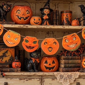 May include: A Halloween garland featuring six die-cut paper pumpkin and cat faces in shades of orange, green, and black, strung together on a natural twine cord. Each cutout displays a vintage-style illustration of a smiling jack-o'-lantern.