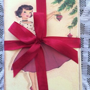 Op de afbeelding: Een set vintage kerstkaarten, vastgebonden met een dieprood satijnen lint. De kaarten tonen een retro illustratie van een vrouw die een kerstboom versiert. De vrouw draagt een polkadot jurk en een bordeauxrode rok. De kaarten zijn vastgebonden met een grote strik.