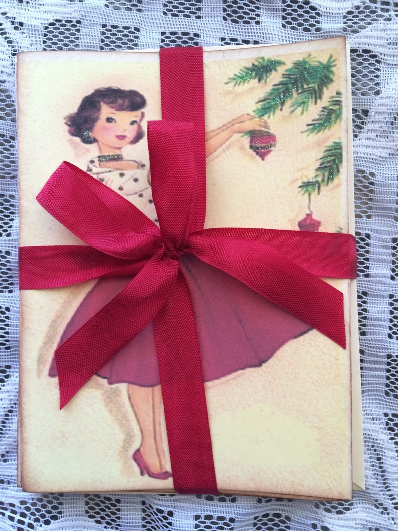 Op de afbeelding: Een set vintage kerstkaarten, vastgebonden met een dieprood satijnen lint. De kaarten tonen een retro illustratie van een vrouw die een kerstboom versiert. De vrouw draagt een polkadot jurk en een bordeauxrode rok. De kaarten zijn vastgebonden met een grote strik.