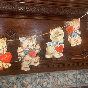 May include: A vintage Valentine's Day garland with five paper cutouts of kittens. Each kitten is wearing a bow and holding a heart-shaped card with a Valentine's Day message. The garland is strung with twine and is hanging on a wooden surface.