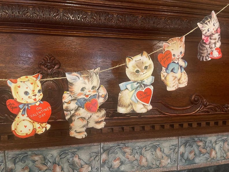 May include: A vintage Valentine's Day garland with five paper cutouts of kittens. Each kitten is wearing a bow and holding a heart-shaped card with a Valentine's Day message. The garland is strung with twine and is hanging on a wooden surface.