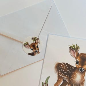 May include: A white envelope with a round sticker featuring a watercolour illustration of a fawn with a holly wreath on its antlers. The sticker is placed on the envelope's flap. A separate card shows a larger watercolour illustration of a fawn with a holly wreath on its antlers.