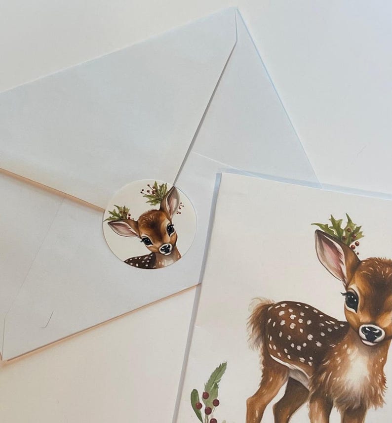 May include: A white envelope with a round sticker featuring a watercolour illustration of a fawn with a holly wreath on its antlers. The sticker is placed on the envelope's flap. A separate card shows a larger watercolour illustration of a fawn with a holly wreath on its antlers.