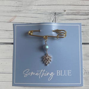 May include: A gold-colored safety pin with a blue bead and a silver-colored leaf charm. The pin is on a light blue card with the words "Something Blue".