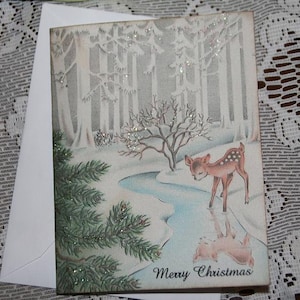 Peut inclure: Carte de No&euml;l vintage repr&eacute;sentant un faon dans une for&ecirc;t enneig&eacute;e avec un ruisseau. La carte pr&eacute;sente un paysage hivernal avec des arbres et l'inscription "Merry Christmas". Comprend une enveloppe blanche.