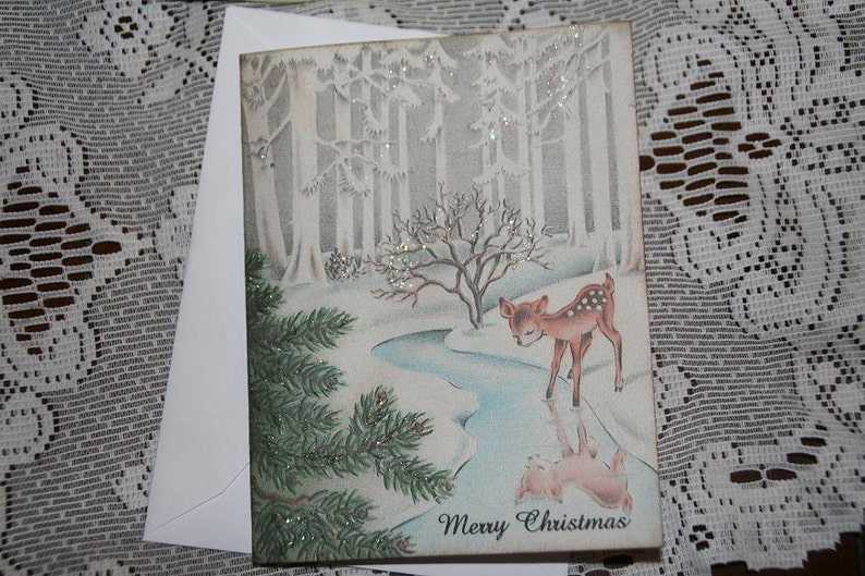 Peut inclure: Carte de No&euml;l vintage repr&eacute;sentant un faon dans une for&ecirc;t enneig&eacute;e avec un ruisseau. La carte pr&eacute;sente un paysage hivernal avec des arbres et l'inscription "Merry Christmas". Comprend une enveloppe blanche.