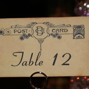 May include: A vintage-style table number card with the words "POST-CARD" and "Table 12" printed on it. The card has an ornate design in blue and is held by a silver metal ring. The background is blurred with bokeh lights.