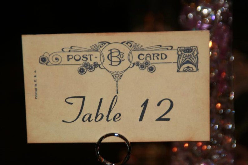 May include: A vintage-style table number card with the words "POST-CARD" and "Table 12" printed on it. The card has an ornate design in blue and is held by a silver metal ring. The background is blurred with bokeh lights.