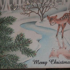 Peut inclure: Carte de No&euml;l vintage avec un faon tachet&eacute; pr&egrave;s d'un cours d'eau. L'illustration comprend un paysage enneig&eacute;, des branches de conif&egrave;res et le texte "Merry Christmas" en &eacute;criture cursive.