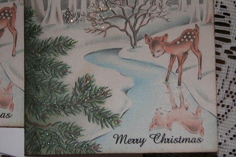 Peut inclure: Carte de No&euml;l vintage avec un faon tachet&eacute; pr&egrave;s d'un cours d'eau. L'illustration comprend un paysage enneig&eacute;, des branches de conif&egrave;res et le texte "Merry Christmas" en &eacute;criture cursive.