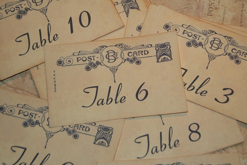 May include: Vintage-style table cards with numbers and the word "Table" in elegant script. Each card features a decorative border with the word "Postcard" and a stamp design. The cards are printed on aged paper.