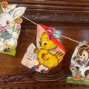 May include: A vintage Easter banner with three colorful paper cutouts. The first cutout features a white bunny with a blue bow tie and the words "Hello!" The second cutout features a yellow chick wearing a red hat and holding a basket. The third cutout features a brown bunny wearing a blue bow tie and holding a basket of eggs. The banner is decorated with flowers and greenery.