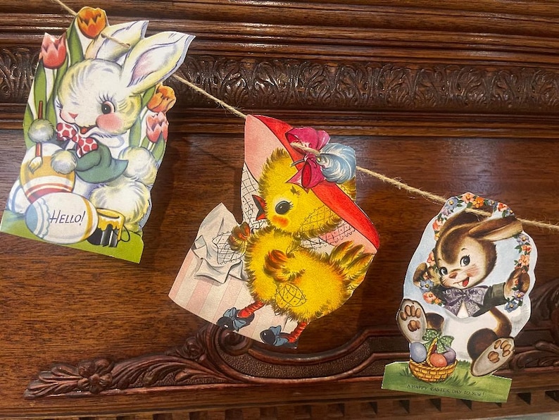 May include: A vintage Easter banner with three colorful paper cutouts. The first cutout features a white bunny with a blue bow tie and the words "Hello!" The second cutout features a yellow chick wearing a red hat and holding a basket. The third cutout features a brown bunny wearing a blue bow tie and holding a basket of eggs. The banner is decorated with flowers and greenery.