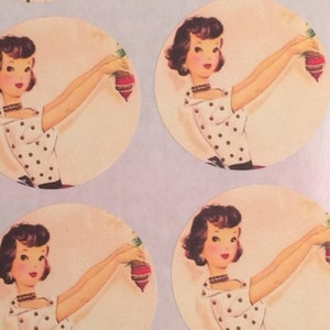 Op de afbeelding: Ronde stickers met een retro-illustratie van een vrouw in een witte polkadotjurk die een rode en groene ornament vasthoudt. De vrouw heeft donker haar en draagt een ketting. De stickers hebben een zacht, vintage kleurenpalet.