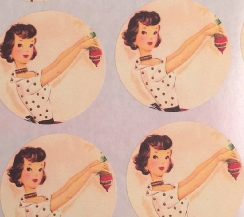Op de afbeelding: Ronde stickers met een retro-illustratie van een vrouw in een witte polkadotjurk die een rode en groene ornament vasthoudt. De vrouw heeft donker haar en draagt een ketting. De stickers hebben een zacht, vintage kleurenpalet.