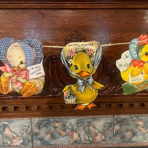 May include: A garland of three Easter-themed ducklings with blue and white gingham bonnets. The center duckling has a blue bow and the text "Happy Easter" above it. The duckling on the left has the text "For Baby's Very First Easter". The duckling on the right has the text "Easter Greetings".