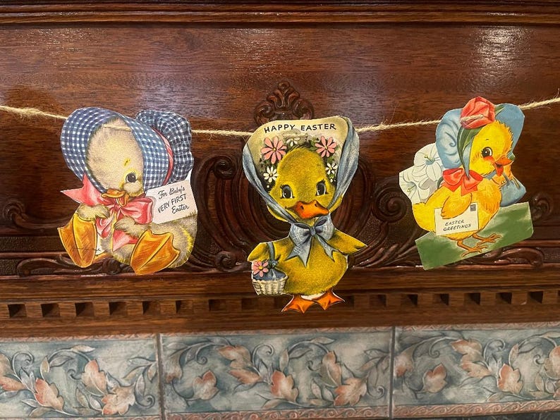 May include: A garland of three Easter-themed ducklings with blue and white gingham bonnets. The center duckling has a blue bow and the text "Happy Easter" above it. The duckling on the left has the text "For Baby's Very First Easter". The duckling on the right has the text "Easter Greetings".