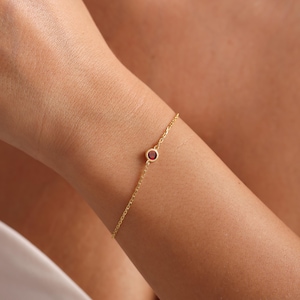 May include: A delicate gold chain bracelet with a single red gemstone.