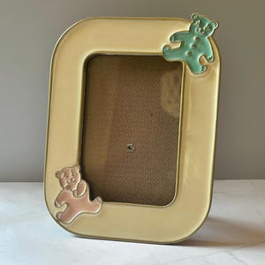 May include: A rectangular photo frame with a cream-colored border and gold trim. Two teddy bear embellishments, one pink and one teal, are on the frame. The frame is designed for a small photo.