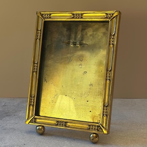 May include: A rectangular brass picture frame with a metallic gold finish. The frame features a decorative border with repeating oval and dot patterns. It stands upright on two small, spherical brass feet at the bottom corners.