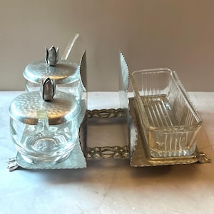 May include: A vintage serving set with a silver-toned metal frame. The set includes two glass jars with hammered metal lids and flower-shaped finials, a glass butter dish, and a serving utensil. The set is designed for serving condiments.