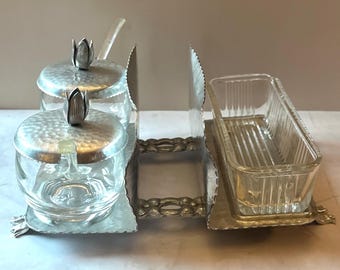 May include: A vintage serving set with a silver-toned metal frame. The set includes two glass jars with hammered metal lids and flower-shaped finials, a glass butter dish, and a serving utensil. The set is designed for serving condiments.