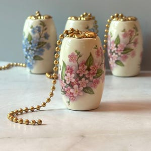 May include: Four decorative ceramic light pull chains with floral designs. The pulls are off-white with gold accents and feature hand-painted flowers in pink and blue. Each pull has a gold ball chain.