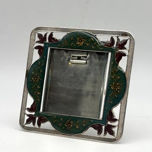 May include: Square silver photo frame with a green and gold floral design. The frame has a decorative border with leaf-shaped accents in burgundy. The center is a silver metal plate.
