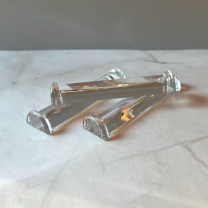 May include: Three clear glass candle holders with a rectangular body and a curved base. The candle holders are lying on a white marble surface. The glass is transparent, allowing light to pass through.