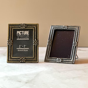 May include: Two small, decorative picture frames. One frame is bronze-colored, the other is silver-toned. Both frames have a floral design and the text "PICTURE THIS" and "2" x 2" (5cm x 5cm)" is visible on one frame.