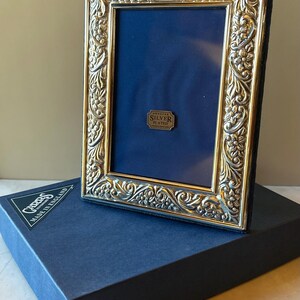 May include: A silver-plated picture frame with an ornate floral design. The frame has a dark blue backing and a small tag that reads "Genuine Silver Plated". The frame is on top of a dark blue box with the text "Made in England".