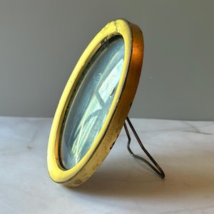 May include: A vintage, oval-shaped, gold-colored picture frame with a stand. The frame has a slightly distressed appearance, with visible wear and tear on the gold finish. The glass is clear, and the frame is angled slightly.