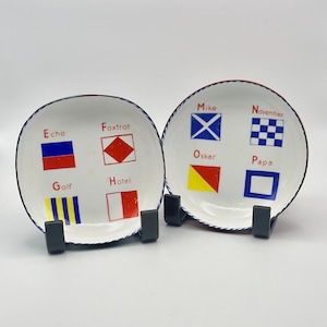May include: Two white ceramic plates with nautical signal flags and names. One plate has "Echo", "Foxtrot", "Golf", and "Hotel". The other plate has "Mike", "November", "Oskar", and "Papa". Each plate is on a black stand.