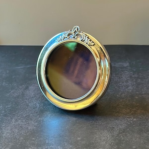 May include: A circular silver-toned metal picture frame with a polished finish. The frame features a decorative embossed leaf motif at the top center and a fine rope-like texture bordering the inner circular opening for a photograph.