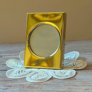 May include: A rectangular gold-toned metal picture frame featuring a circular center opening. The frame has a smooth, reflective surface and a clean, simple design, providing a polished appearance for displaying a single photograph or image.
