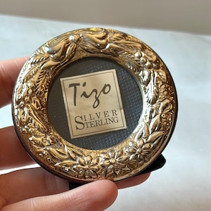 May include: A small, round, silver-colored picture frame with an ornate floral and ribbon design. The frame holds a small, dark-colored backing with a label that reads "Tizo SILVER STERLING". The frame is held by a hand.