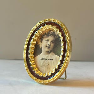 May include: An oval-shaped picture frame with a metallic gold finish and a scalloped inner border. The frame contains a vintage-style portrait of a child and features the text MADE IN TAIWAN printed clearly across the lower portion of the image.
