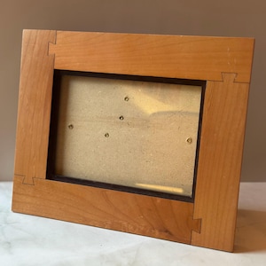 May include: A rectangular wooden picture frame with a light brown finish. The frame has a dark brown interior border and is assembled with interlocking joints at the corners. The frame is designed to hold a photograph or artwork.