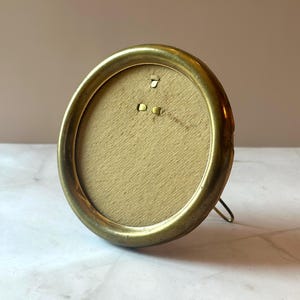 May include: An oval-shaped, antique brass picture frame. The frame has a simple design with a slightly tarnished finish, and a small metal stand on the back. The interior backing is a light tan color.