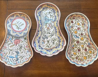 May include: Three decorative ceramic spoon rests with floral and bird designs. Each rest has a unique pattern in shades of blue, yellow, red, and green, with scalloped edges. The rests are displayed on a wooden surface.
