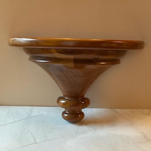 May include: A wooden wall shelf with a rounded top and a decorative, turned support. The shelf is made of a warm-toned wood, with visible grain patterns. The shelf is designed to be mounted on a wall and is suitable for displaying decorative items.