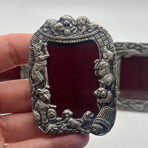 May include: A silver-coloured picture frame with a rectangular opening and a decorative border featuring sculpted puppies. The frame has a dark red interior. The frame is a unique decorative item.