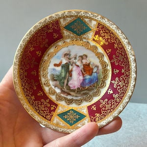 May include: A small decorative porcelain plate featuring a central circular painting of three figures in flowing garments. The border is decorated with deep red panels, gold filigree patterns, and teal diamond shapes with gold accents.