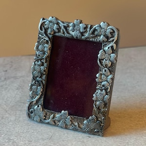 May include: A rectangular picture frame made of a metallic silver-toned material with a dark maroon velvet backing. The frame features a raised, embossed floral pattern of dogwood blossoms and leaves covering the entire border surface.