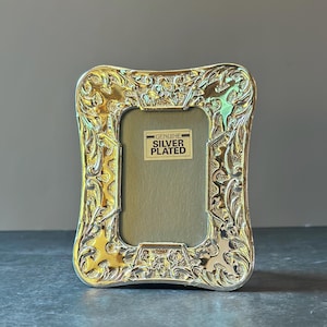May include: A silver-plated picture frame with a rectangular opening and an ornate, embossed floral border. A rectangular label in the center reads "GENUINE SILVER PLATED" in black text on a white background, set against a gray backing.